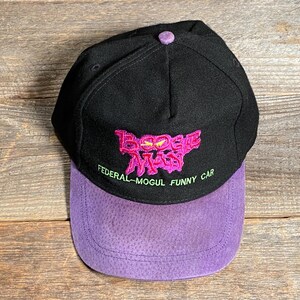 May include: Black baseball cap with a purple suede bill. The cap features embroidered text in pink and green that reads "Boogie Man" and "Federal-Mogul Funny Car". The cap has a button on top.