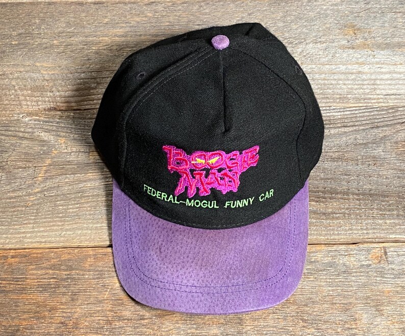 May include: Black baseball cap with a purple suede bill. The cap features embroidered text in pink and green that reads "Boogie Man" and "Federal-Mogul Funny Car". The cap has a button on top.