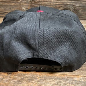 May include: Black baseball cap with a burgundy accent. The cap features a flat peak and an adjustable snapback fastening. The hat is made from a sturdy material and is suitable for everyday wear.