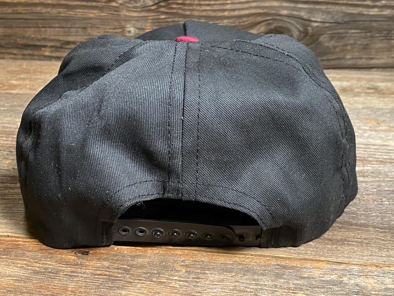 May include: Black baseball cap with a burgundy accent. The cap features a flat peak and an adjustable snapback fastening. The hat is made from a sturdy material and is suitable for everyday wear.