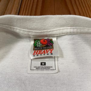 Puede incluir: Primer plano de una camiseta blanca con cuello de canal&eacute;. La etiqueta dice "FRUIT OF THE LOOM HEAVY COTTON" con un gr&aacute;fico de frutas. Debajo del logo hay una etiqueta con la talla "M" y el texto "100% COTTON PRESHRUNK MADE IN HONDURAS."