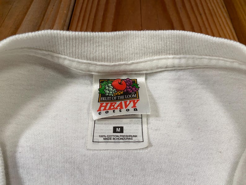 Puede incluir: Primer plano de una camiseta blanca con cuello de canal&eacute;. La etiqueta dice "FRUIT OF THE LOOM HEAVY COTTON" con un gr&aacute;fico de frutas. Debajo del logo hay una etiqueta con la talla "M" y el texto "100% COTTON PRESHRUNK MADE IN HONDURAS."