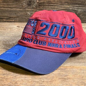 May include: A red baseball cap with a blue brim featuring embroidered text that reads 2000, Hot Rod Association, and Auto Club NHRA Finals in blue thread. The cap includes a small blue American flag graphic and a circular logo on the front panel.