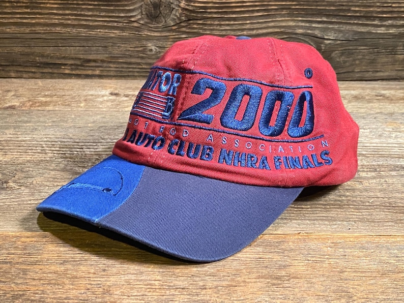 May include: A red baseball cap with a blue brim featuring embroidered text that reads 2000, Hot Rod Association, and Auto Club NHRA Finals in blue thread. The cap includes a small blue American flag graphic and a circular logo on the front panel.