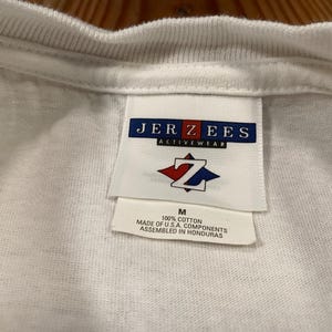 May include: Close-up of a white t-shirt collar and tag. The tag reads "JERZEES ACTIVEWEAR" with a red and blue logo. Below the logo is the size "M" and text indicating the shirt is 100% cotton and assembled in Honduras.