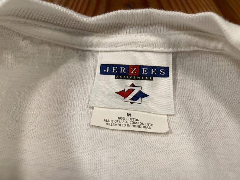 May include: Close-up of a white t-shirt collar and tag. The tag reads "JERZEES ACTIVEWEAR" with a red and blue logo. Below the logo is the size "M" and text indicating the shirt is 100% cotton and assembled in Honduras.