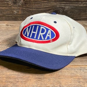 Op de afbeelding: Een witte baseballcap met een blauw vizier. De cap heeft de tekst "NHRA" in blauwe letters in een rood en blauw ovaal. De hoed ligt op een houten oppervlak.