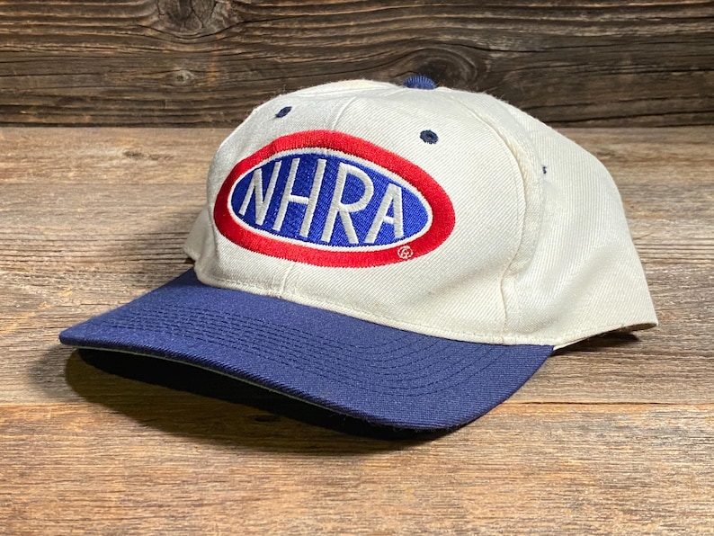 Op de afbeelding: Een witte baseballcap met een blauw vizier. De cap heeft de tekst "NHRA" in blauwe letters in een rood en blauw ovaal. De hoed ligt op een houten oppervlak.