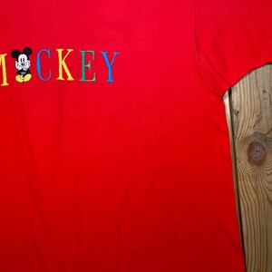 May include: A bright red short-sleeved t-shirt featuring the embroidered text MOCKEY in multicolored letters. The letter O is replaced by a small, embroidered graphic of a cartoon mouse with black ears, a white face, and yellow shorts.