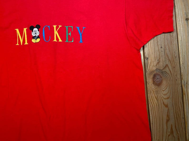 May include: A bright red short-sleeved t-shirt featuring the embroidered text MOCKEY in multicolored letters. The letter O is replaced by a small, embroidered graphic of a cartoon mouse with black ears, a white face, and yellow shorts.