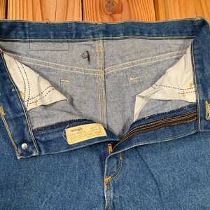 May include: Close-up of blue denim jeans with the zipper partially open. The jeans have a button closure and a small tag with the brand name "Wrangler". The interior pockets are visible, and the stitching is in yellow.