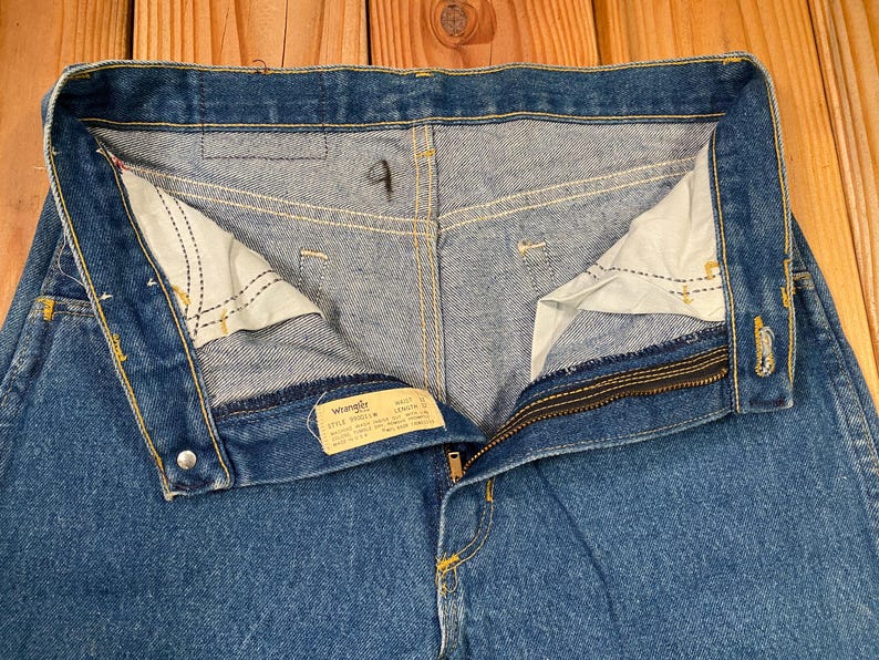 May include: Close-up of blue denim jeans with the zipper partially open. The jeans have a button closure and a small tag with the brand name "Wrangler". The interior pockets are visible, and the stitching is in yellow.