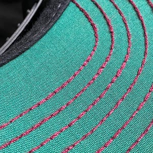 May include: Close-up of a baseball cap's interior. The cap features a teal green fabric with curved, burgundy-coloured stitching. The brim's edge is lined with black fabric.
