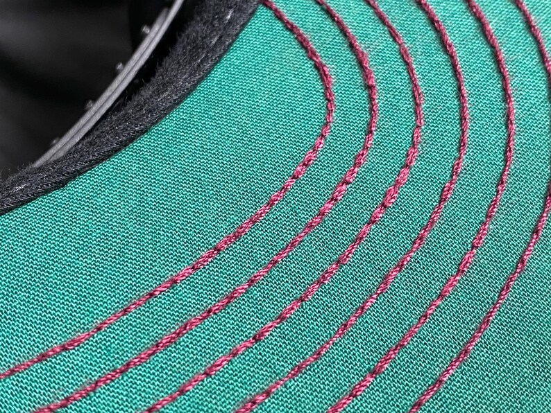 May include: Close-up of a baseball cap's interior. The cap features a teal green fabric with curved, burgundy-coloured stitching. The brim's edge is lined with black fabric.