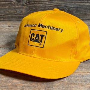 May include: A bright yellow baseball cap with a flat brim. The cap features the text "Johnson Machinery" above a black and yellow "CAT" logo.