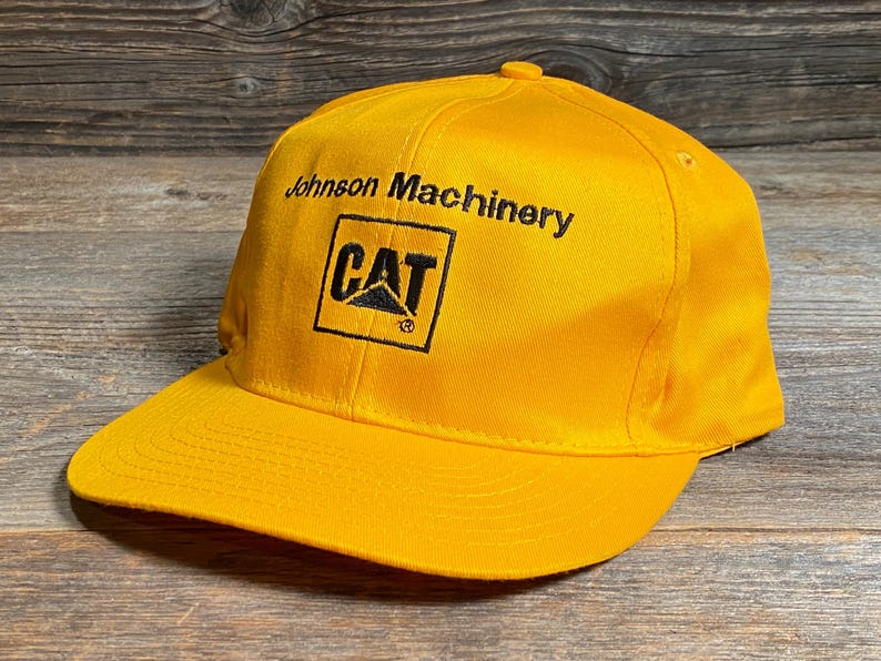 May include: A bright yellow baseball cap with a flat brim. The cap features the text "Johnson Machinery" above a black and yellow "CAT" logo.