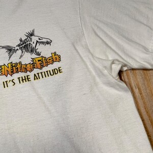 Puede incluir: Camiseta blanca con un gr&aacute;fico negro y naranja de un esqueleto de pez, las palabras "Nitro Fish" y "It's the Attitude" en naranja y amarillo. La camiseta tiene mangas cortas y cuello redondo.