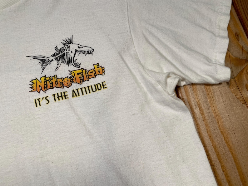 Puede incluir: Camiseta blanca con un gr&aacute;fico negro y naranja de un esqueleto de pez, las palabras "Nitro Fish" y "It's the Attitude" en naranja y amarillo. La camiseta tiene mangas cortas y cuello redondo.