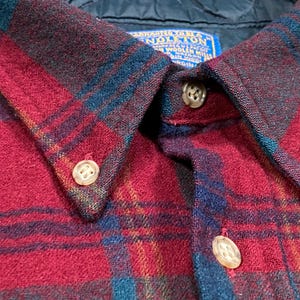 May include: Close-up of a red, blue, and green plaid button-down shirt. The shirt features a classic collar with three visible buttons. The fabric has a textured appearance, and a blue label is visible near the collar.