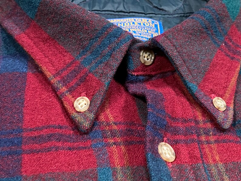 May include: Close-up of a red, blue, and green plaid button-down shirt. The shirt features a classic collar with three visible buttons. The fabric has a textured appearance, and a blue label is visible near the collar.