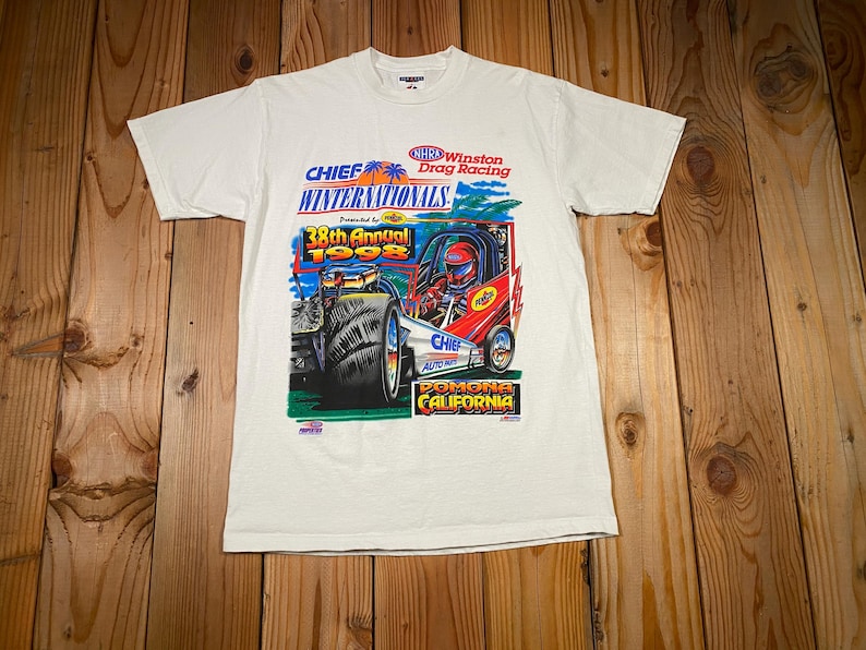 May include: White t-shirt featuring a colorful graphic of a drag racing car with the text "CHIEF WINTERNATIONALS 1998" and "POMONA CALIFORNIA". The shirt has short sleeves and a crew neck.