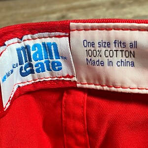 Op de afbeelding: De binnenkant van een felrode honkbalpet met twee witte stoffen labels. Het linkerlabel toont het blauwe Main Gate-logo. Het rechterlabel bevat de tekst: One size fits all, 100% COTTON, Made in china, gedrukt in donkerblauw en zwarte letters.