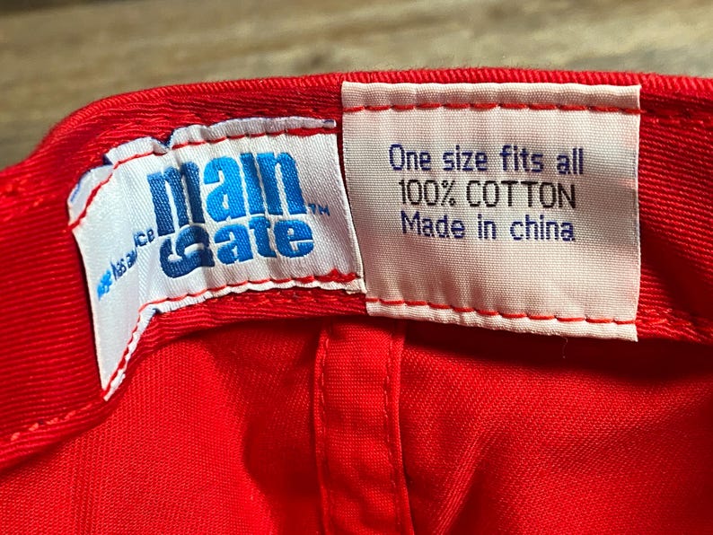 Op de afbeelding: De binnenkant van een felrode honkbalpet met twee witte stoffen labels. Het linkerlabel toont het blauwe Main Gate-logo. Het rechterlabel bevat de tekst: One size fits all, 100% COTTON, Made in china, gedrukt in donkerblauw en zwarte letters.