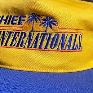 May include: A yellow baseball cap with a blue brim. The cap features the text "CHIEF WINTERNATIONALS" in blue embroidery with a palm tree and sunset design.