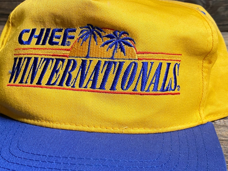 May include: A yellow baseball cap with a blue brim. The cap features the text "CHIEF WINTERNATIONALS" in blue embroidery with a palm tree and sunset design.