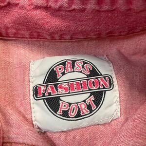 Puede incluir: Una prenda de mezclilla roja con una etiqueta de tela rectangular blanca cosida en el cuello. La etiqueta muestra un emblema circular negro con el texto PASS FASHION PORT en letras rojas, con la palabra FASHION centrada en una barra negra.