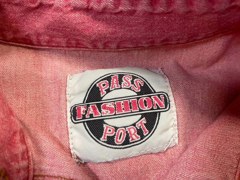 Puede incluir: Una prenda de mezclilla roja con una etiqueta de tela rectangular blanca cosida en el cuello. La etiqueta muestra un emblema circular negro con el texto PASS FASHION PORT en letras rojas, con la palabra FASHION centrada en una barra negra.