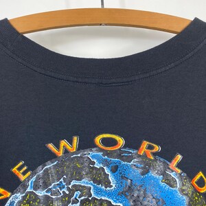 May include: Black t-shirt with the words "NEW WORLD" in orange and yellow lettering above a blue and green globe design. The shirt is hanging on a wooden hanger.