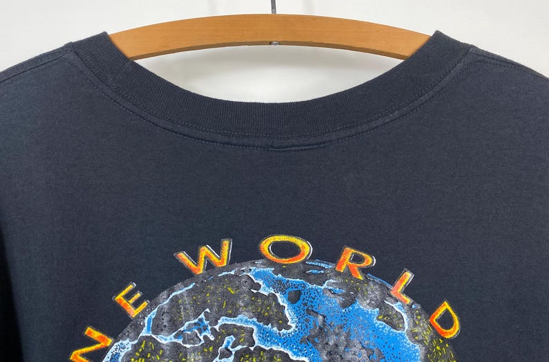 May include: Black t-shirt with the words "NEW WORLD" in orange and yellow lettering above a blue and green globe design. The shirt is hanging on a wooden hanger.