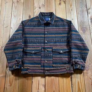 Puede incluir: Una chaqueta de manga larga con un patrón horizontal repetitivo en tonos azul marino, naranja óxido y beige. La chaqueta incluye un cuello estándar, cierre frontal de botones, dos bolsillos grandes con solapa en el pecho y un dobladillo elástico.