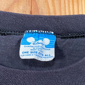 Puede incluir: Una camiseta negra con una etiqueta de cuello rectangular azul y blanca. La etiqueta muestra el logotipo de Disney Character Fashions, el texto Made in USA y la frase One Size Fits All, con instrucciones para ver el reverso para el cuidado.