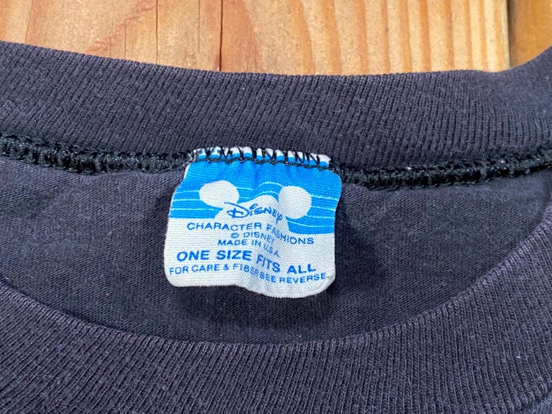 Puede incluir: Una camiseta negra con una etiqueta de cuello rectangular azul y blanca. La etiqueta muestra el logotipo de Disney Character Fashions, el texto Made in USA y la frase One Size Fits All, con instrucciones para ver el reverso para el cuidado.