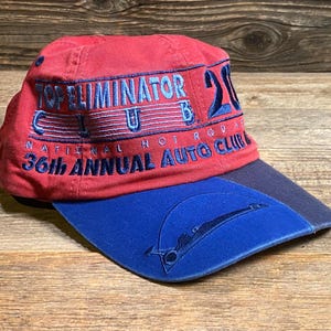 May include: A red baseball cap with a blue brim featuring embroidered text that reads Top Eliminator Club 2000 National Hot Rod Association 36th Annual Auto Club. The blue brim includes a stitched graphic of a drag racing vehicle silhouette.