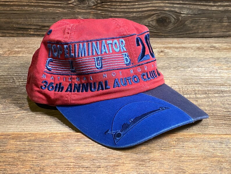 May include: A red baseball cap with a blue brim featuring embroidered text that reads Top Eliminator Club 2000 National Hot Rod Association 36th Annual Auto Club. The blue brim includes a stitched graphic of a drag racing vehicle silhouette.