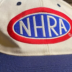 Op de afbeelding: Een cr&egrave;mekleurige baseballpet met een marineblauwe klep. De pet heeft een groot ovaal logo met de letters "NHRA" in wit, omlijnd in rood en tegen een blauwe achtergrond. De pet heeft een klassiek zes-panelen ontwerp.
