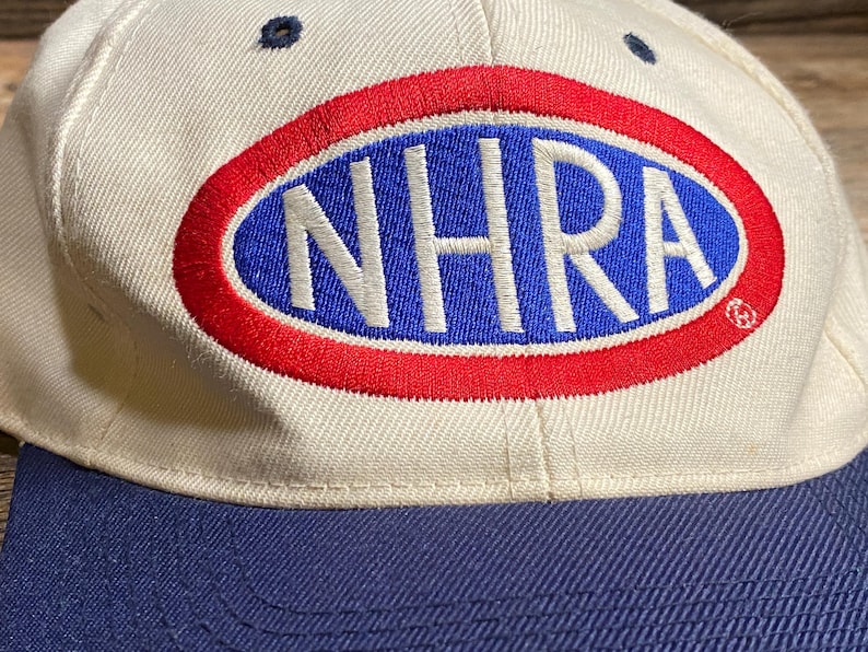 Op de afbeelding: Een cr&egrave;mekleurige baseballpet met een marineblauwe klep. De pet heeft een groot ovaal logo met de letters "NHRA" in wit, omlijnd in rood en tegen een blauwe achtergrond. De pet heeft een klassiek zes-panelen ontwerp.