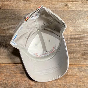 May include: A light grey baseball cap with a white brim and a logo. The interior is white with a logo. The cap is on a wooden surface.
