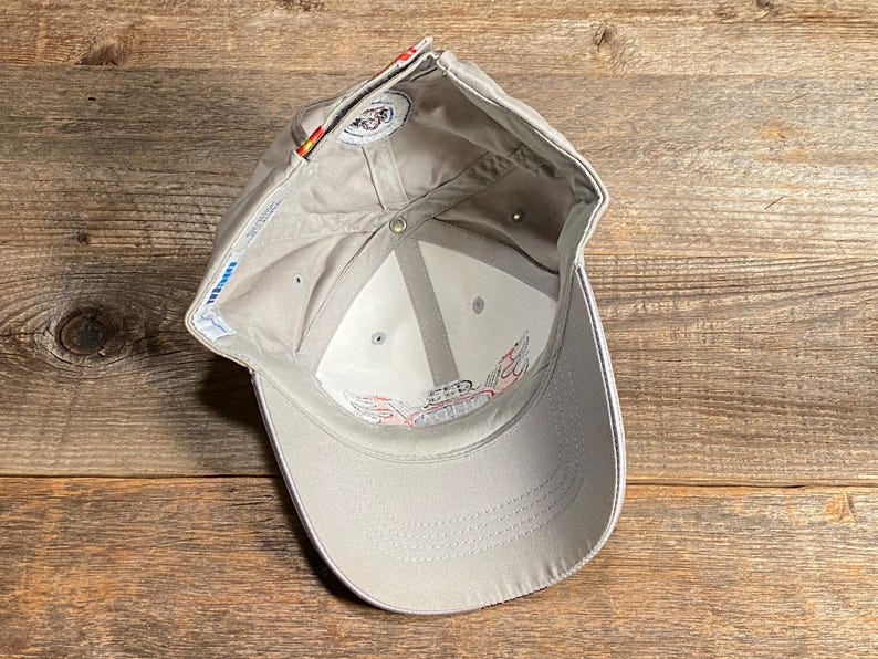 May include: A light grey baseball cap with a white brim and a logo. The interior is white with a logo. The cap is on a wooden surface.