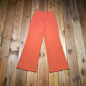 May include: A pair of orange high-waisted pants with a flared leg design. The pants are made of a solid color fabric and have a button and zipper closure. The pants are laid flat on a wooden surface.