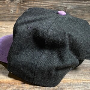 May include: A black baseball cap with a purple brim and a matching purple button on top. The cap is made of a textured fabric and has a classic, structured design.