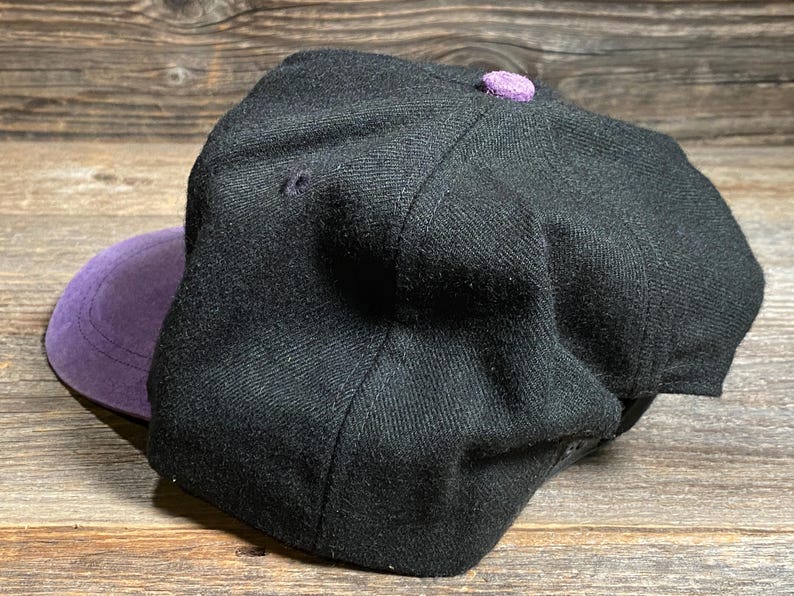 May include: A black baseball cap with a purple brim and a matching purple button on top. The cap is made of a textured fabric and has a classic, structured design.