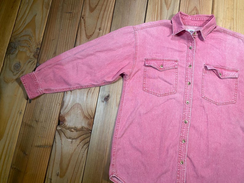Puede incluir: Una camisa de manga larga con botones en tela de mezclilla rosa deste&ntilde;ida. La camisa presenta un cuello est&aacute;ndar, una tapeta de botones frontal completa y dos bolsillos en el pecho con solapas abotonadas. Las mangas tienen pu&ntilde;os abotonados.