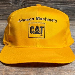 May include: A yellow baseball cap with a curved brim. The cap features the text "Johnson Machinery" above a black square with the "CAT" logo. The hat is made of a durable fabric.