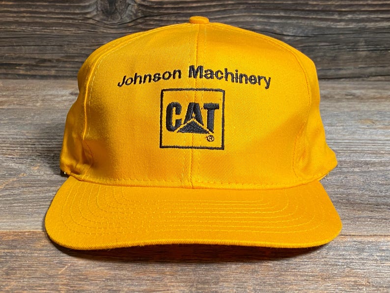 May include: A yellow baseball cap with a curved brim. The cap features the text "Johnson Machinery" above a black square with the "CAT" logo. The hat is made of a durable fabric.