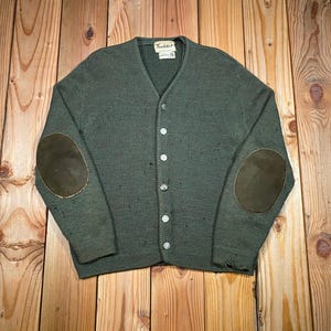 May include: A dark olive green knit cardigan featuring a V-neckline, a front button closure with five light-colored buttons, and contrasting dark brown oval elbow patches on each sleeve. The garment includes a small white brand label at the neck.