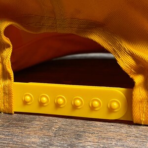 May include: Close-up of a yellow baseball cap, showing the adjustable strap and the underside of the brim. The cap is made of a textured fabric and has a bright yellow color. The adjustable strap is also yellow.